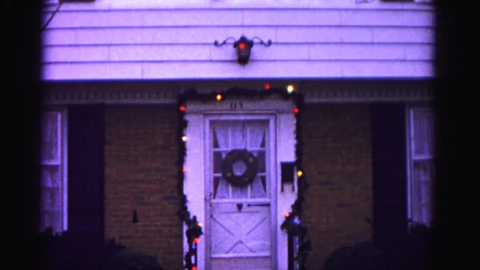 Brick-and-clapboard residential exterior ca. 1962, Hagerstown, Maryland: white door with wreath and colored holiday string lights; purple color shift from aged 8mm reversal stock.