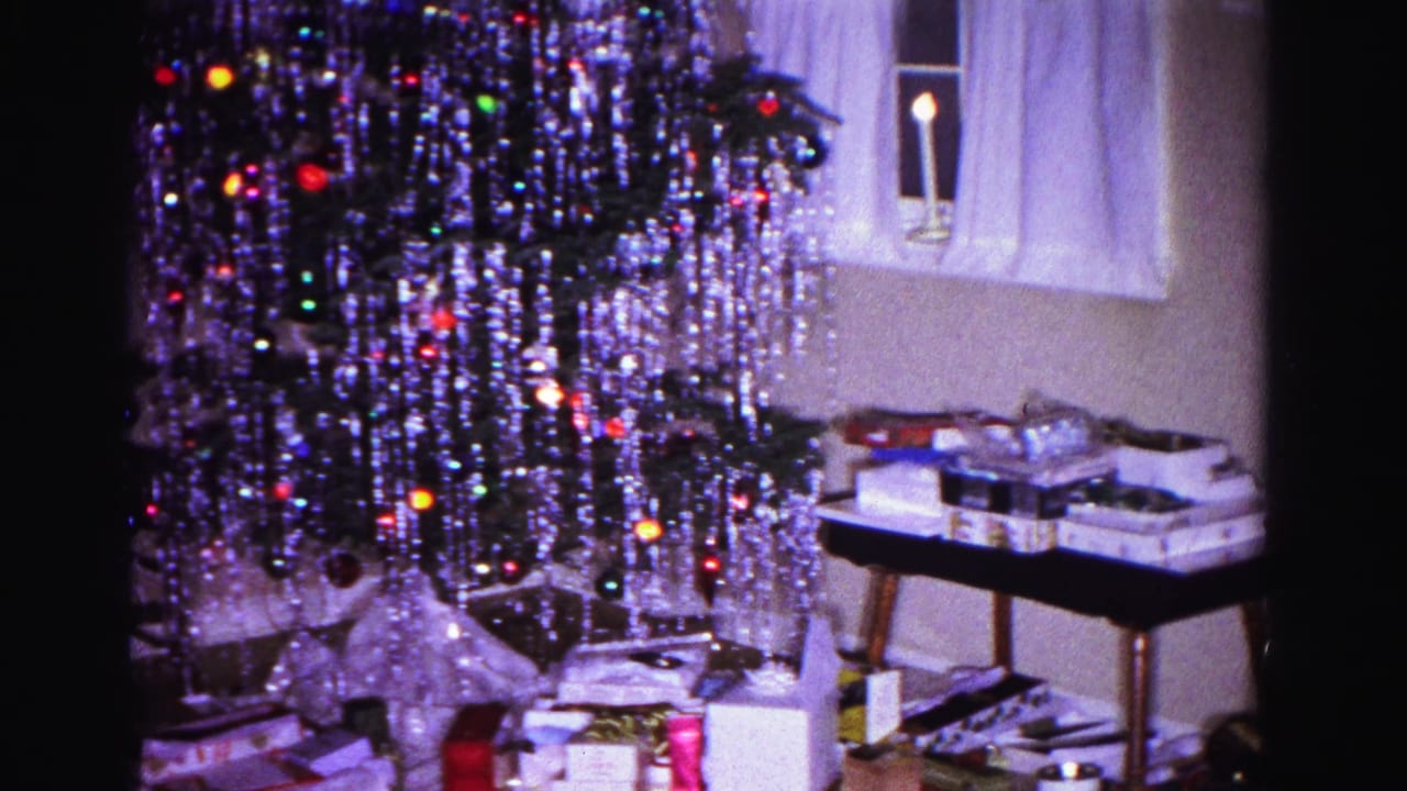 8mm home movie frame from 1962 showing a Christmas tree with heavy silver tinsel and colored lights, wrapped gifts on a table, and a candle in a curtained window, Hagerstown, Maryland