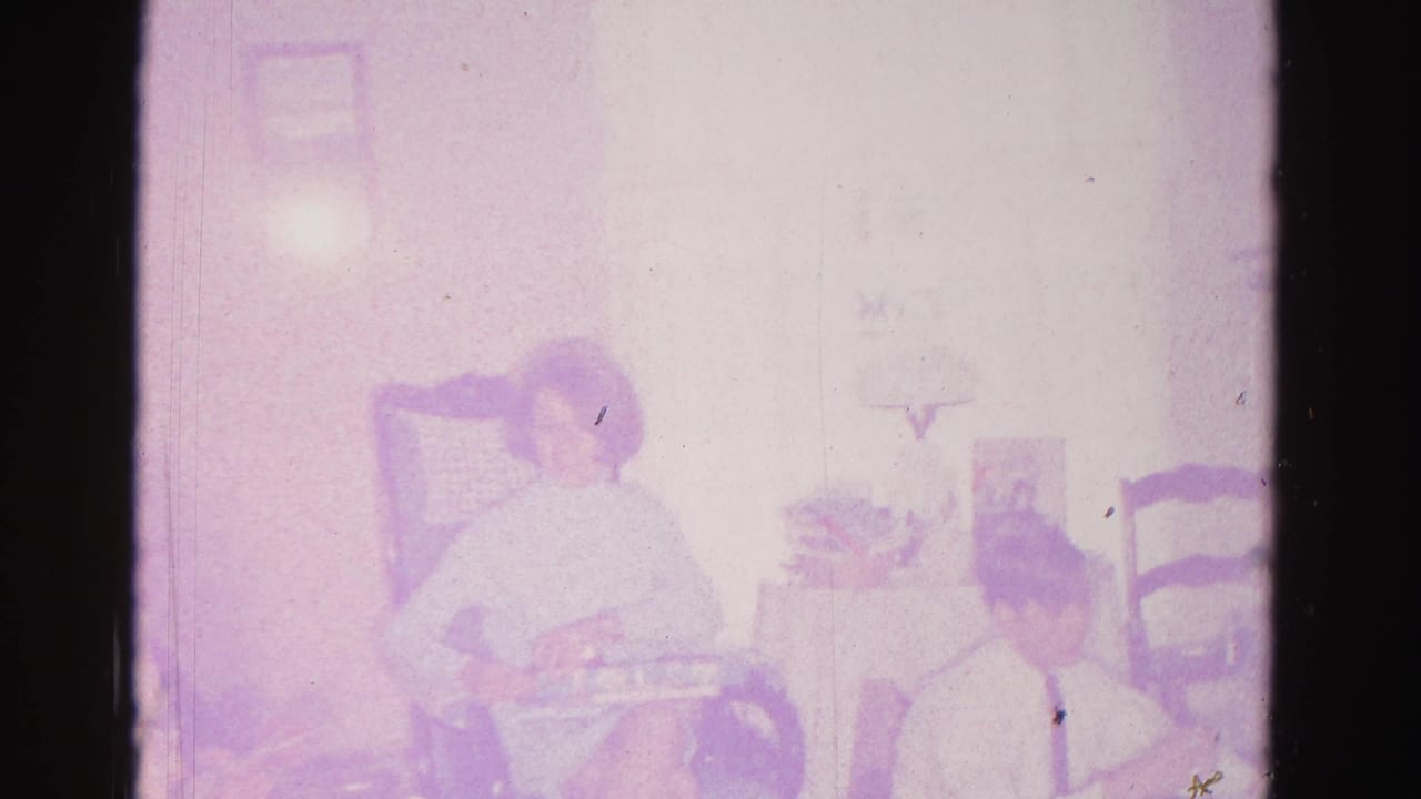 Two adults in what appears to be a 1962 Hagerstown, Maryland living room; one seated in a wooden chair handles a gift box, heavy purple 8mm color cast throughout.