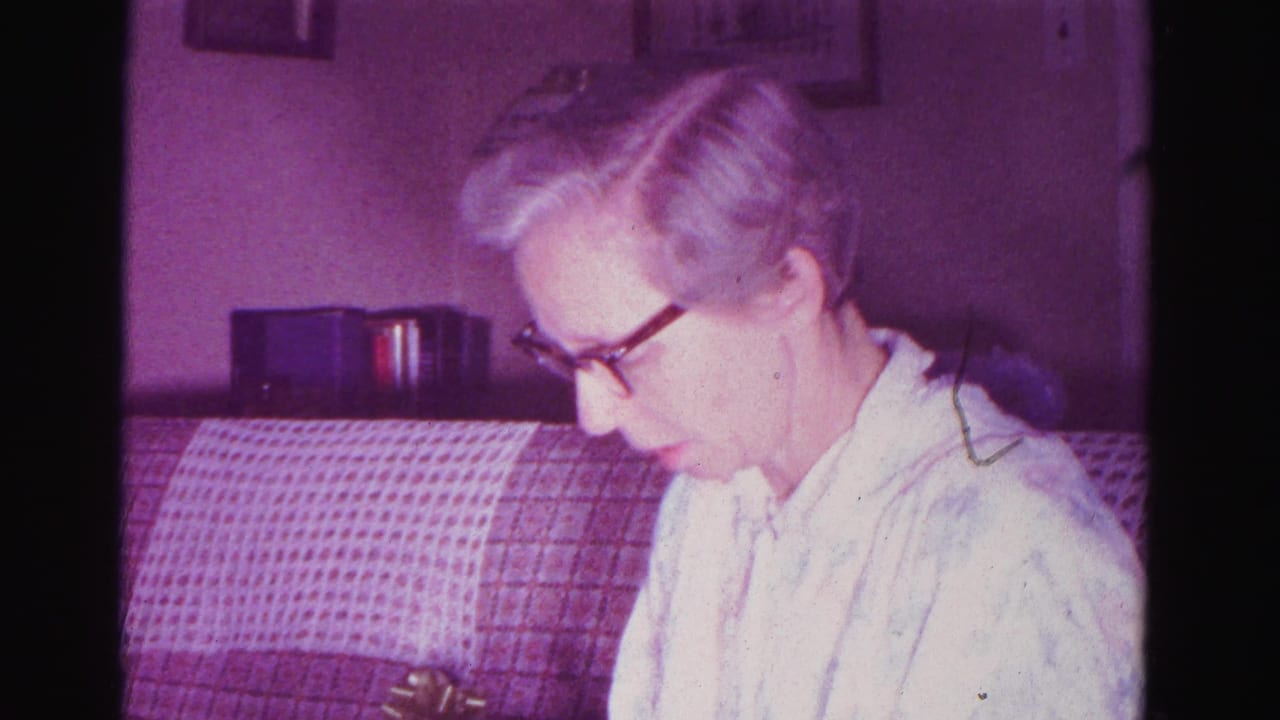 Close-up of an older woman in cat-eye glasses and a light blouse looking downward, seated beside a plaid upholstered sofa.