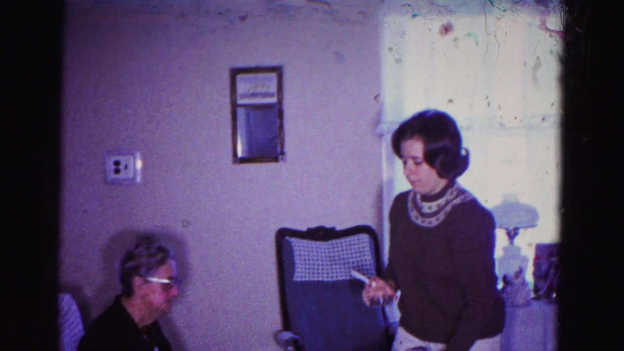Two women in a living room: older woman with glasses seated at left, younger woman in a dark patterned sweater standing at right holding an object.
