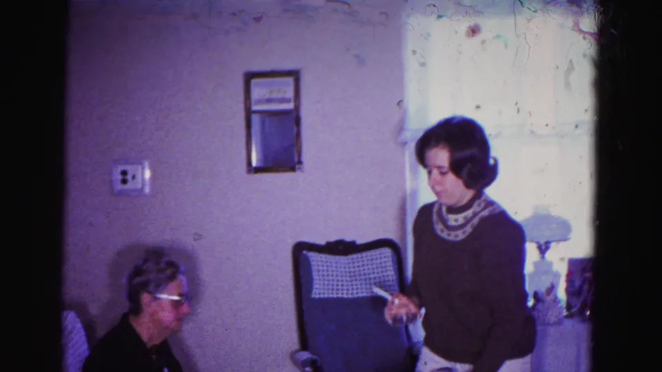 Wider shot of the same 1962 Maryland living room showing an older woman with glasses seated at left and a younger woman in a dark sweater with patterned collar standing at right, holding what may be a card or gift.