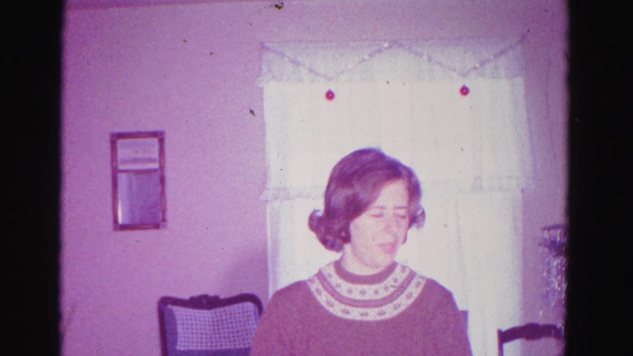 Younger woman in a rust-toned Fair Isle-style patterned sweater near a window with sheer white curtains and a decorative valance; footage consistent with a 1962 Christmas home movie from Hagerstown, Maryland.