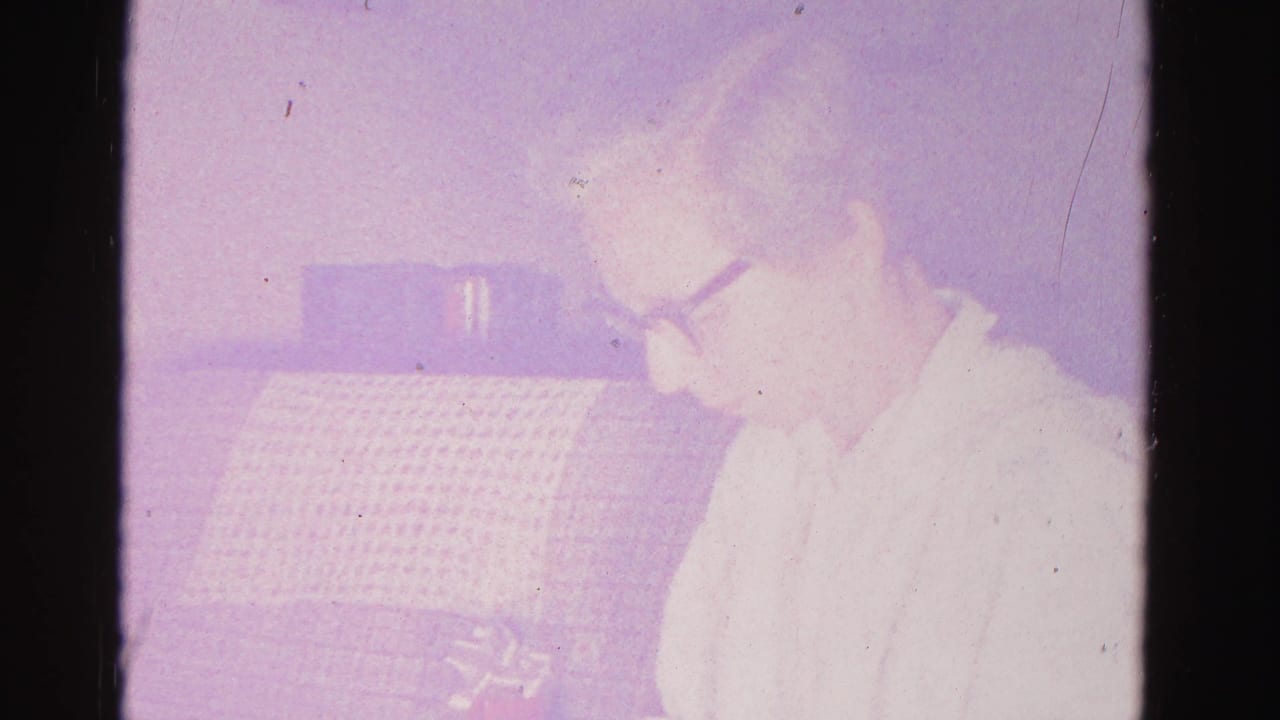 Older woman in dark-framed glasses and a light blouse seen in three-quarter profile looking down toward her hands, grid-textured upholstered couch behind her.