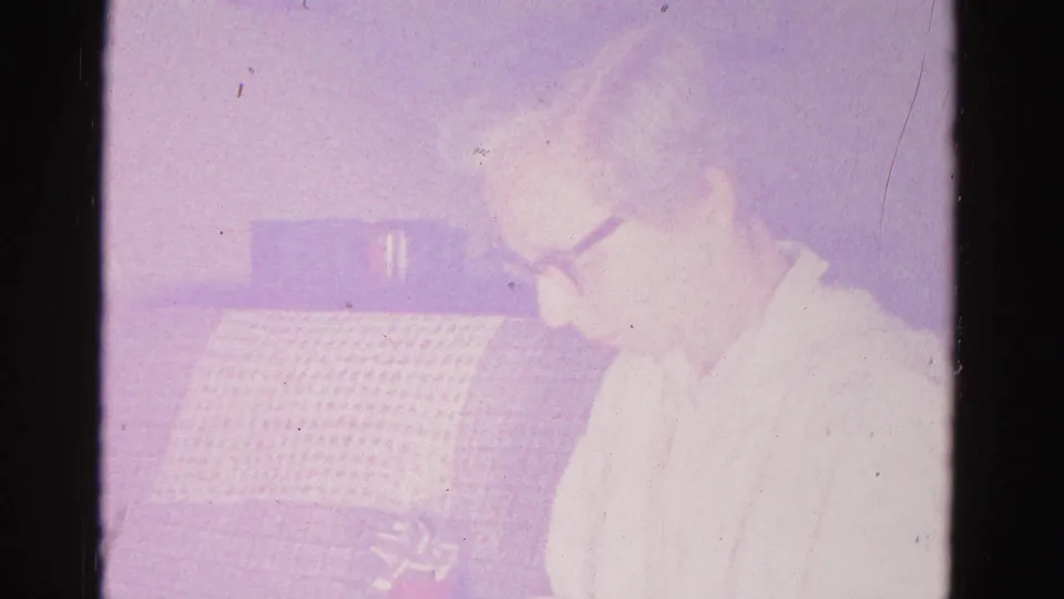 Older woman in early-1960s dark-framed glasses and light blouse examining something in her hands; grid-textured couch behind; 8mm home movie, Hagerstown, Maryland, 1962.