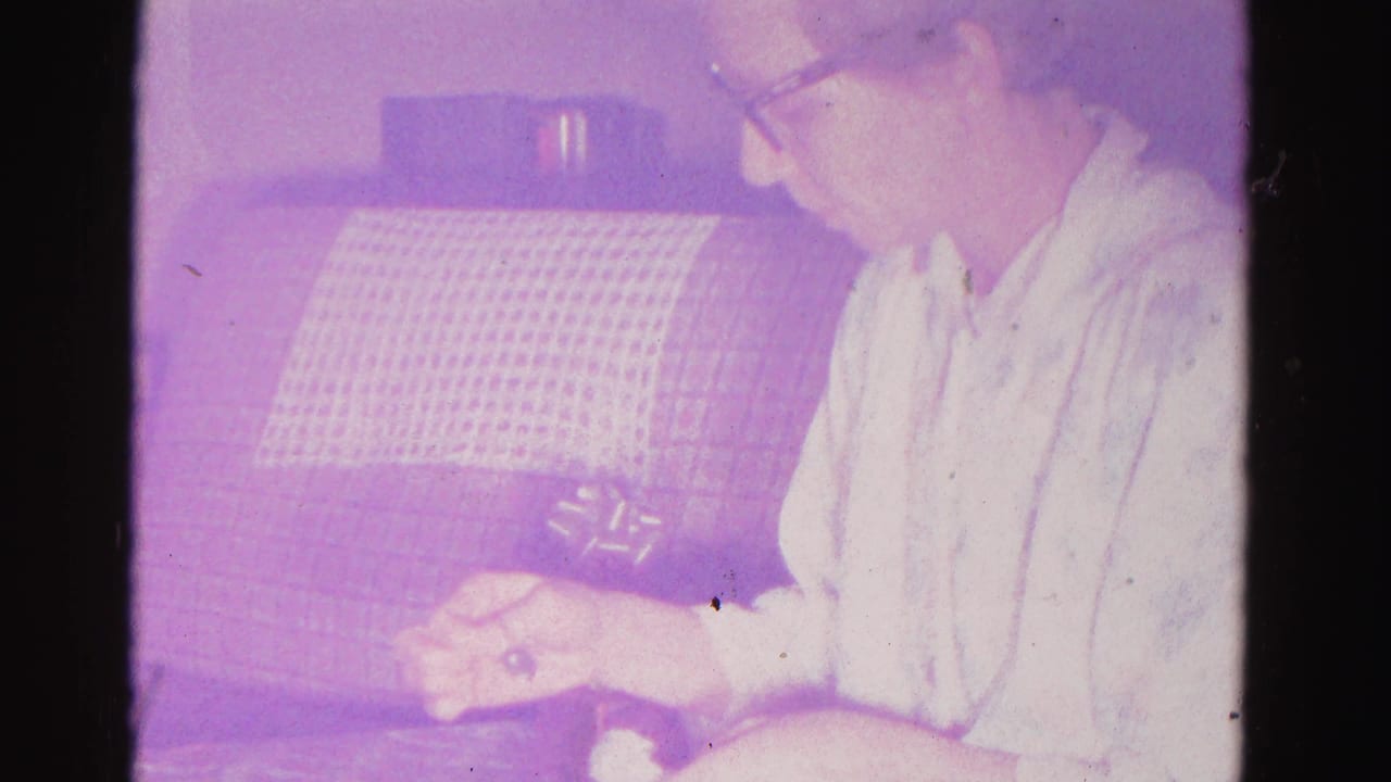 Older woman in dark-framed glasses and light patterned blouse focused on a small object in both hands; grid-textured couch behind; 8mm home movie, Hagerstown, Maryland, 1962.