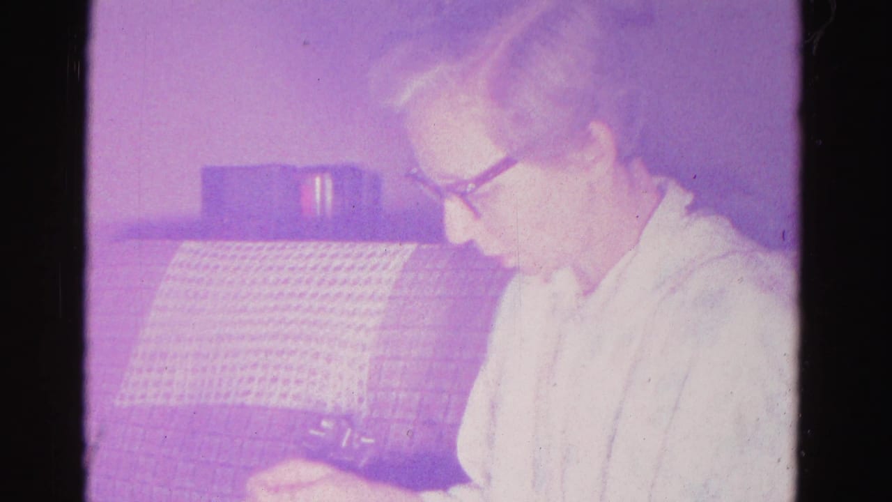 Close-profile view of an older woman in dark-framed glasses examining something in her hands; grid-textured couch behind; 8mm home movie, Hagerstown, Maryland, 1962.