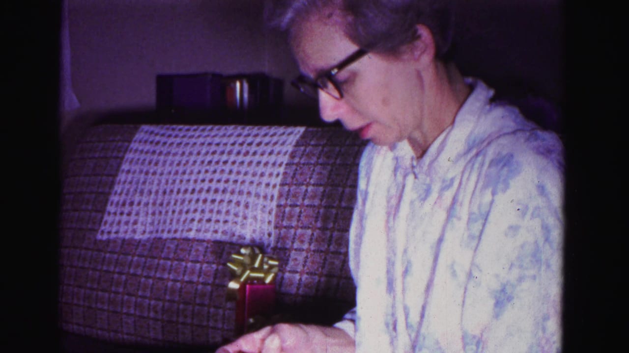An older woman wearing dark-framed glasses examines a small red gift box with a gold bow in a domestic interior; 8mm film, Hagerstown, Maryland, 1962.