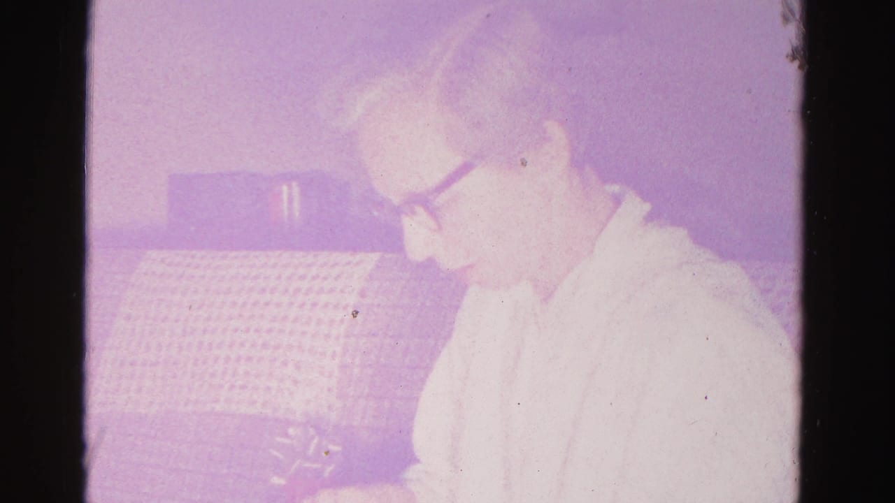 An older woman with dark-framed glasses gazes downward in a domestic interior; 8mm film with prominent purple cast and visible grain, Hagerstown, Maryland, c.1962.