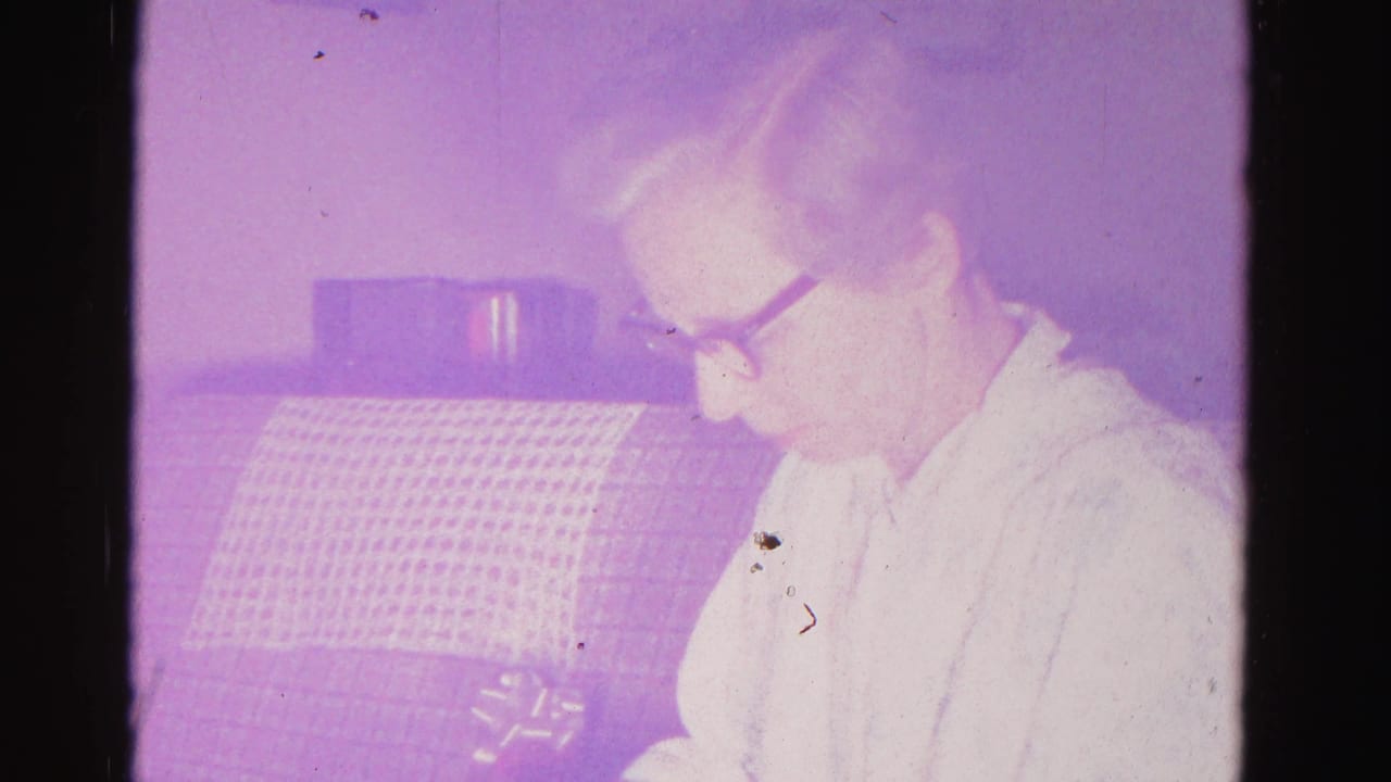An older woman wearing dark-framed glasses holds an object in a domestic interior; aged 8mm film with purple cast and film dust, Hagerstown, Maryland, c.1962.