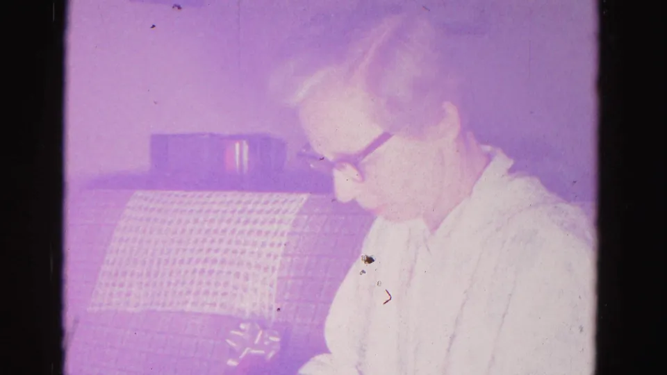 An older woman wearing dark-framed glasses holds an object in a domestic interior; aged 8mm film with purple cast and film dust, Hagerstown, Maryland, c.1962.
