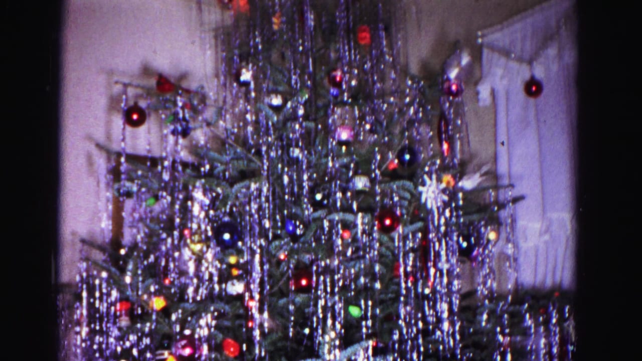 Wider view of the Christmas tree showing tinsel and multicolor glass ornaments, with a curtained window partially visible at upper right.