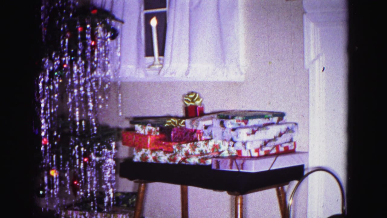 Wrapped Christmas gifts with ribbon bow on a folding tray stand, windowsill candle and white curtains in background, tinsel-draped tree above; 8mm home movie, Hagerstown Maryland, 1962.