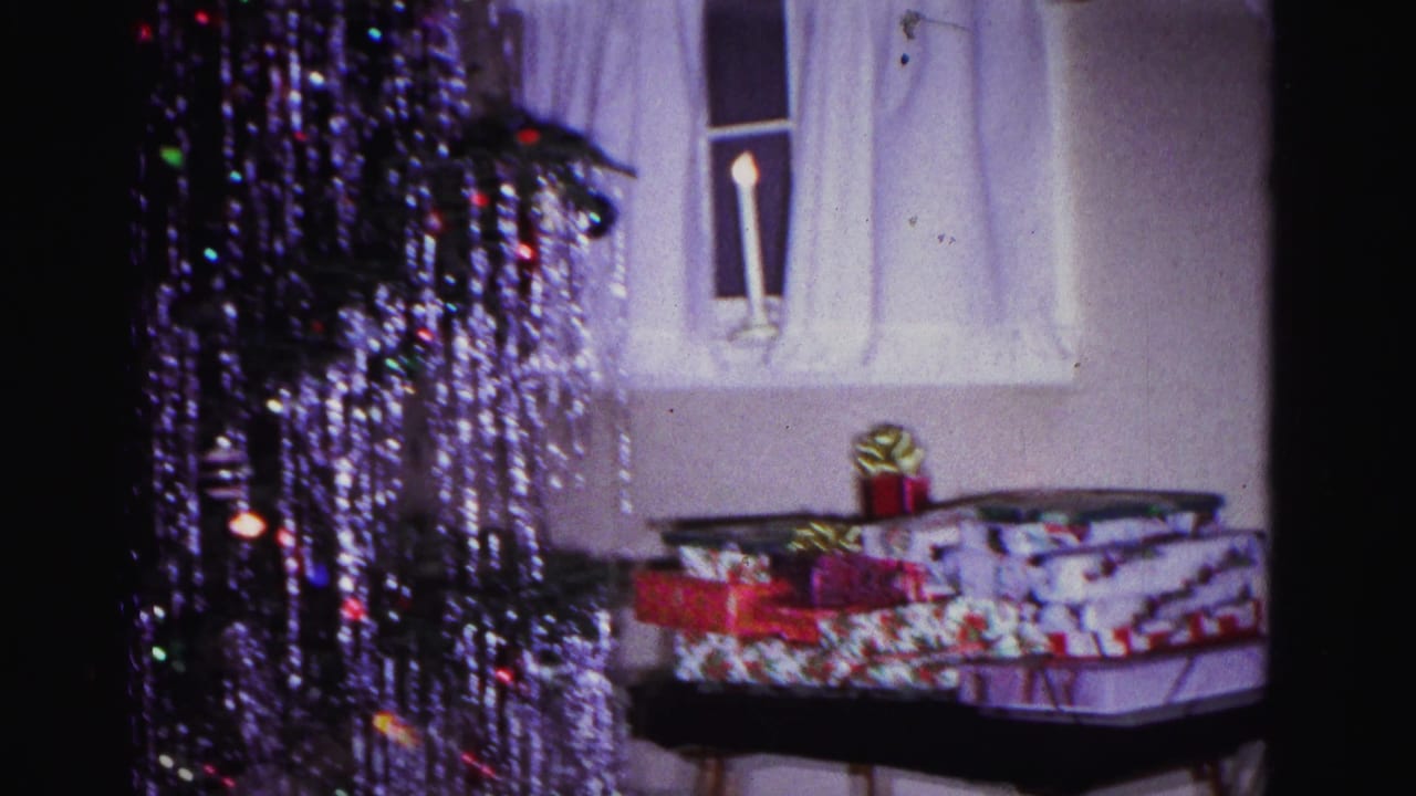 Holiday-wrapped gifts and ribbon bow on a folding tray table, curtained window and lit windowsill candle in domestic interior, tinsel tree above; 8mm home movie, Hagerstown Maryland, 1962.
