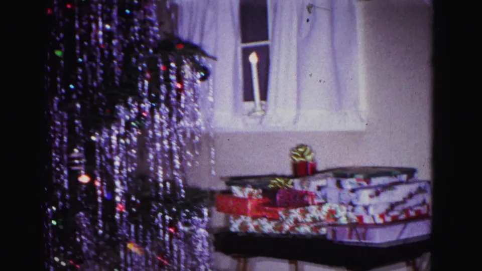 Holiday-wrapped gifts and ribbon bow on a folding tray table, curtained window and lit windowsill candle in domestic interior, tinsel tree above; 8mm home movie, Hagerstown Maryland, 1962.
