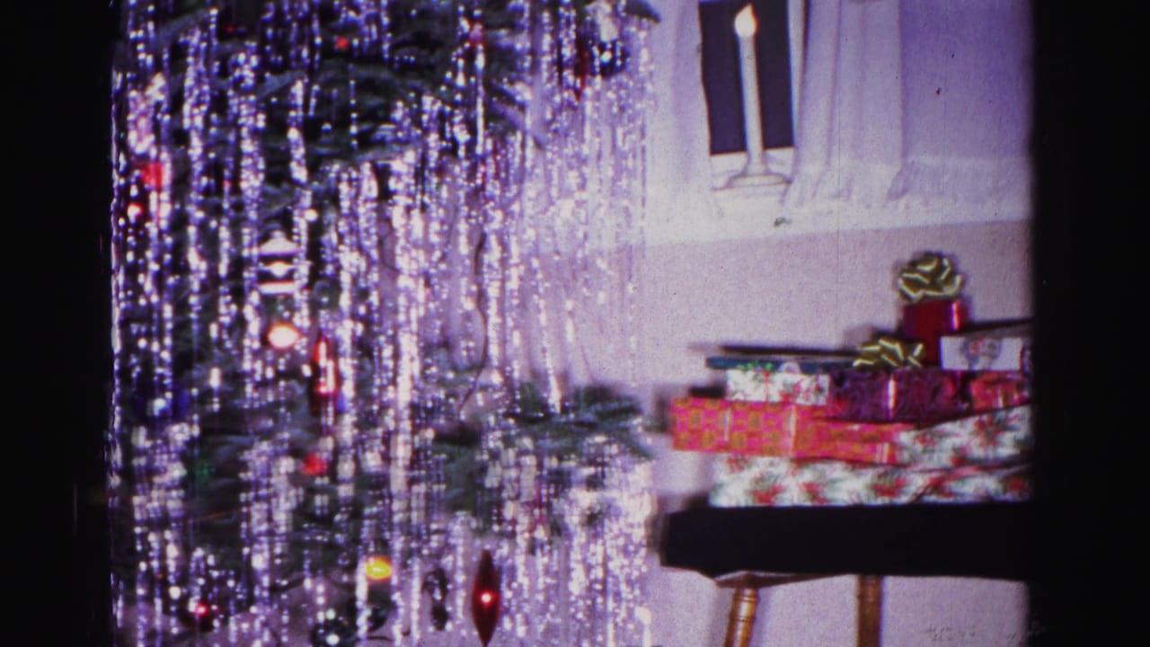 Wooden side table stacked with wrapped gifts beside a tinsel-draped Christmas tree; residential interior with window in background.
