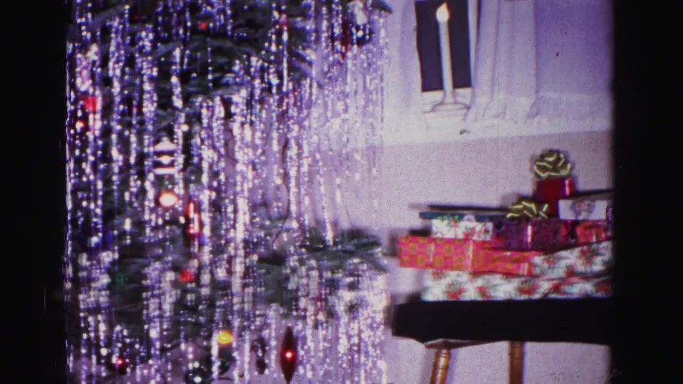 8mm frame, circa 1962, showing a wooden side table piled with festively wrapped gifts next to a tinsel-draped Christmas tree in a private residential interior.