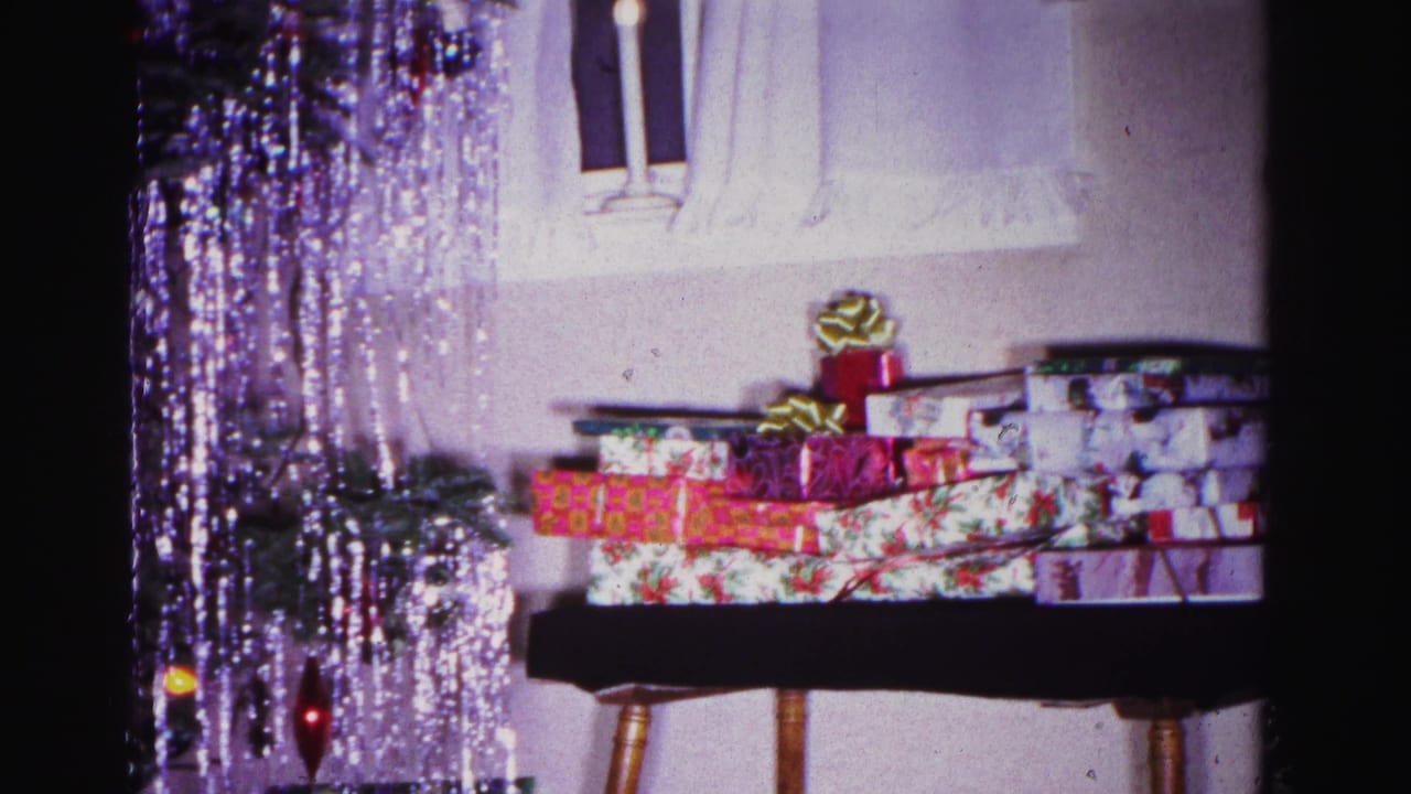 8mm home movie frame, circa 1962, showing a stack of wrapped holiday gifts with a gold bow on a wooden table; silver tinsel from the Christmas tree hangs in the upper foreground.