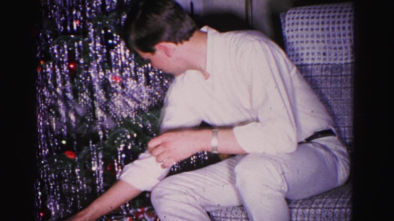 Man in white shirt adjusts ornaments near the base of a heavily tinsel-draped Christmas tree in what appears to be a 1962 Hagerstown, Maryland home.