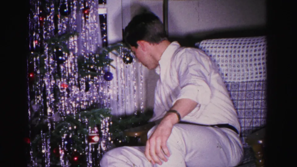A man in a white shirt and wristwatch leans toward a Christmas tree dense with silver tinsel, his face visible in profile, in an apparent 1962 home movie.