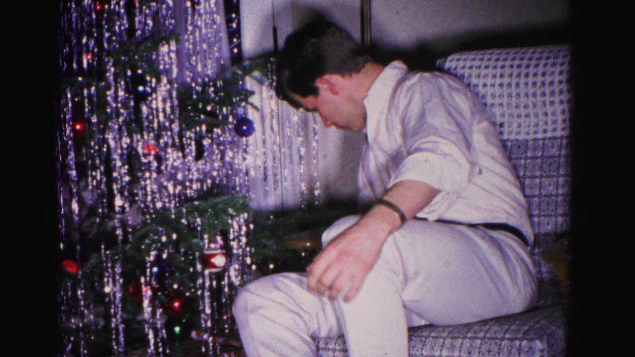 A man bends forward near the base of a tinsel-draped Christmas tree in an early 1960s home movie interior, apparently adjusting lower decorations.