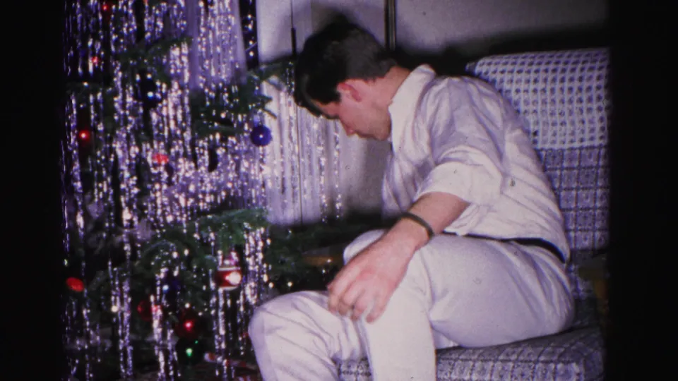 A man bends forward near the base of a tinsel-draped Christmas tree in an early 1960s home movie interior, apparently adjusting lower decorations.