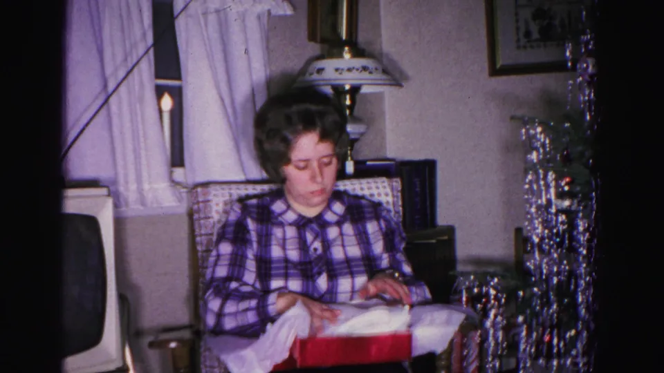 Young woman in plaid shirt unwrapping a red gift box, circa 1962, Hagerstown Maryland; silver tinsel Christmas tree and CRT television visible in background.