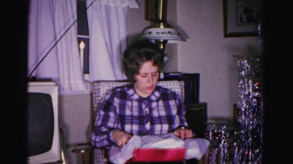 Woman in plaid shirt pausing during Christmas gift opening, circa 1962, Hagerstown Maryland; tinsel tree, CRT television, and curtained window with candle in background.