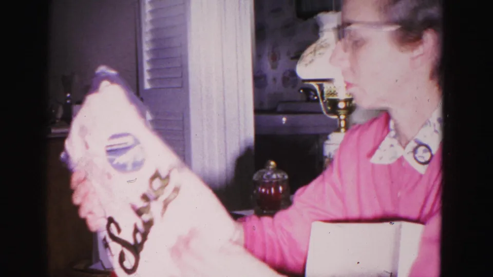 Woman in pink sweater with white collar examining a branded package at Christmas, circa 1962, Hagerstown Maryland; brass candlestick and interior shelving visible.