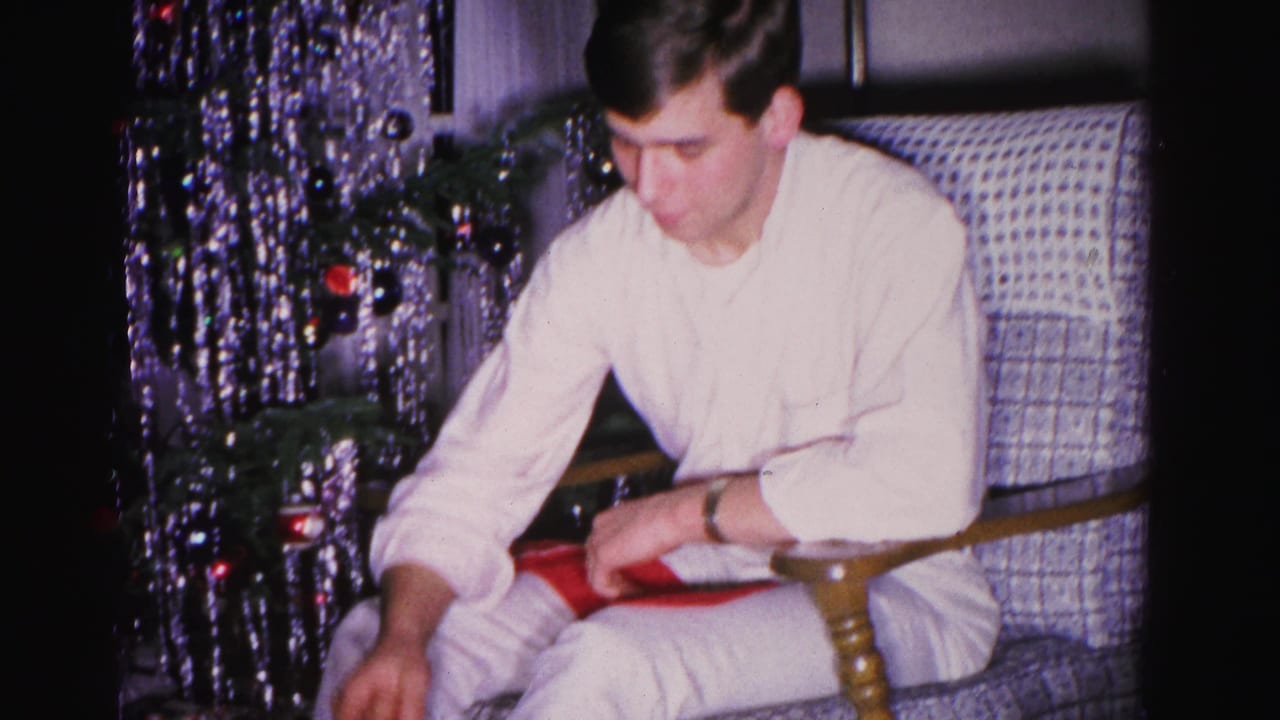 Young man in white shirt unwraps a red gift in a decorated living room; Christmas tree with silver tinsel at left; 8mm home movie, Hagerstown, Maryland, 1962.