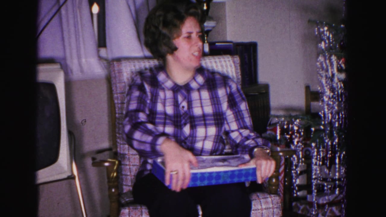 Woman in purple-and-white plaid blouse seated in an armchair holding a blue gift box, silver tinsel decoration visible at right.