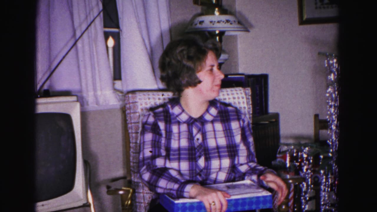 Wider shot of woman in plaid blouse with blue gift box; boxy vintage television set and silver tinsel decorations visible; 8mm home movie, Hagerstown, Maryland, 1962.