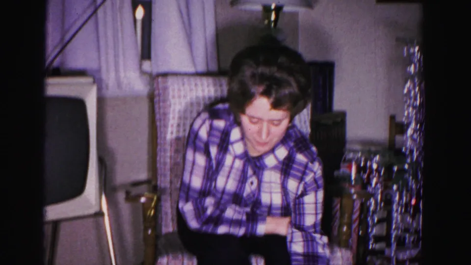 Woman in plaid blouse examines blue gift box; vintage television and silver tinsel visible in background; 8mm home movie, Hagerstown, Maryland, 1962.