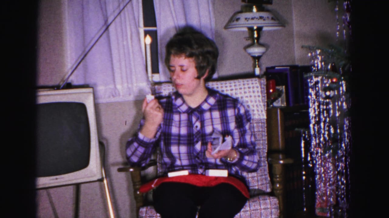 Young woman in blue plaid shirt seated in a patterned armchair, raising something to her mouth, with a vintage CRT television visible at left.