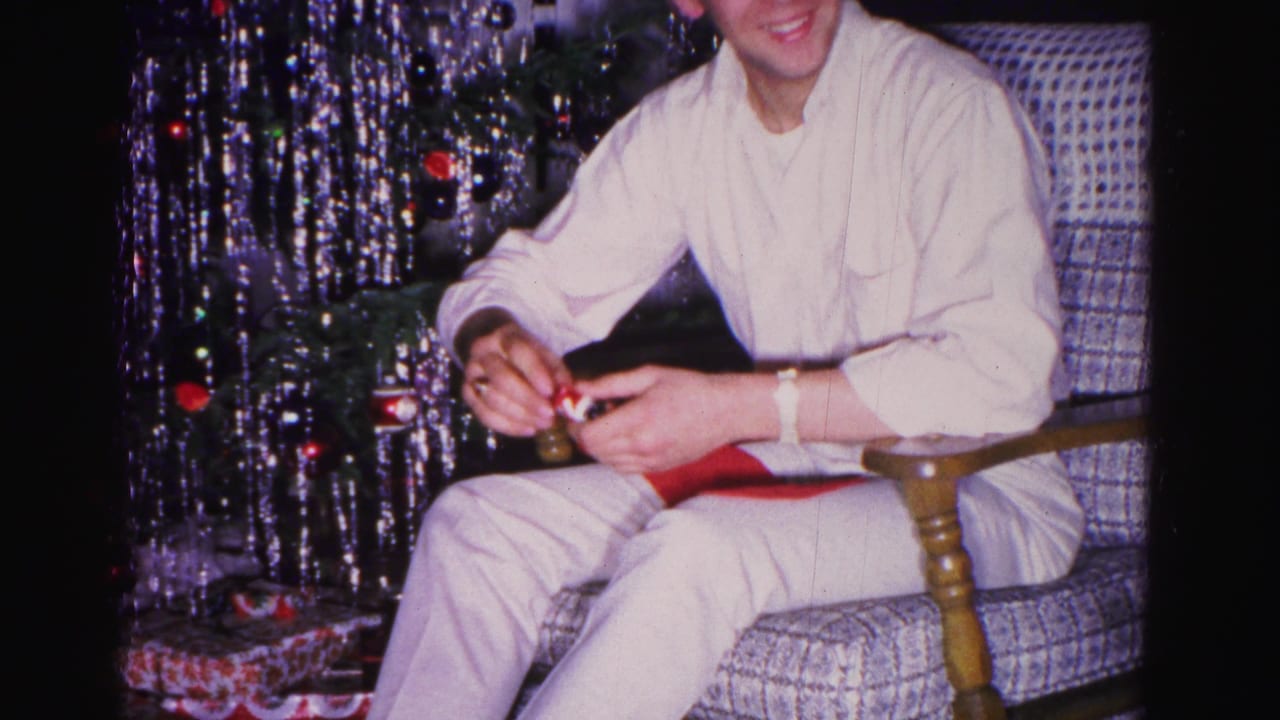 Young man in white long-sleeve shirt smiling as he opens a gift in a patterned armchair, with a metallic aluminum tinsel Christmas tree visible — 8mm home movie, circa 1962.