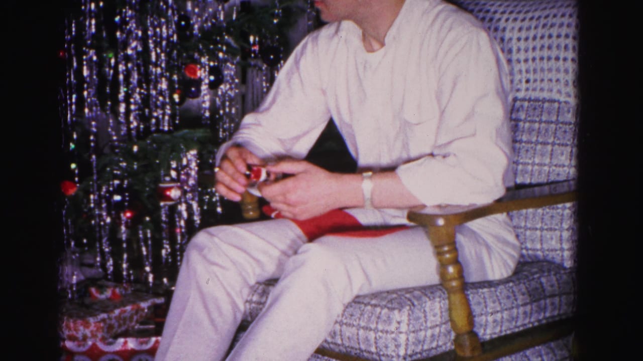 Young man in white shirt and light trousers examining a gift in his lap while seated in a checkered armchair, metallic Christmas tree behind — 8mm home movie, circa 1962.