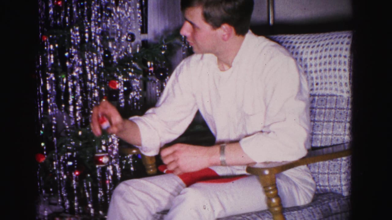 Young man in white shirt smiling as he holds an unwrapped item, patterned armchair and metallic tinsel Christmas tree behind — 8mm home movie, circa 1962.