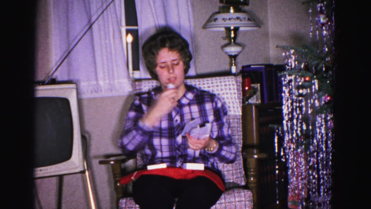 Woman in purple plaid shirt examines an unwrapped gift item in a 1962 Hagerstown living room; vintage boxy television, table lamp, and silver tinsel garland visible in background.