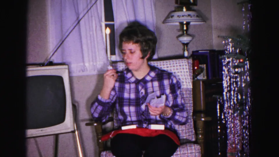 Woman in purple plaid shirt reads or inspects a gift item in an early-1960s Hagerstown living room; silver tinsel garland, vintage television, and table lamp visible in background.