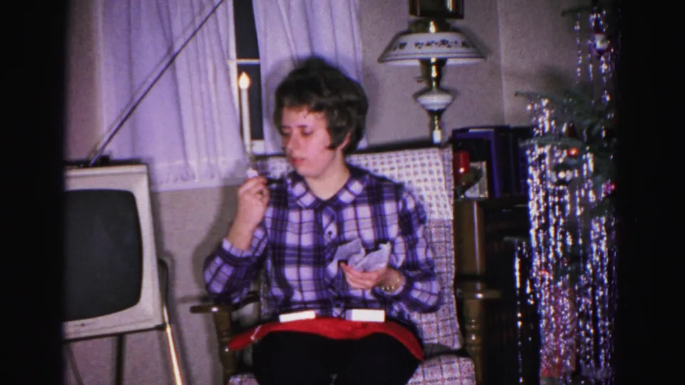 Woman in purple plaid shirt continues inspecting her Christmas gift in a 1962 Hagerstown living room; boxy vintage television, silver tinsel garland, and table lamp visible.
