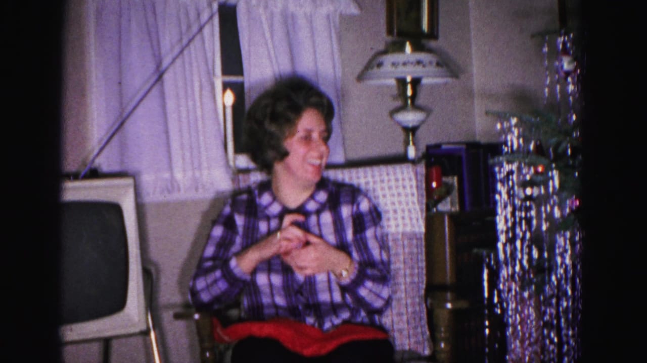 Woman in plaid blouse holding a red item, seated in a mid-century living room with a CRT television and tinsel decoration behind her.