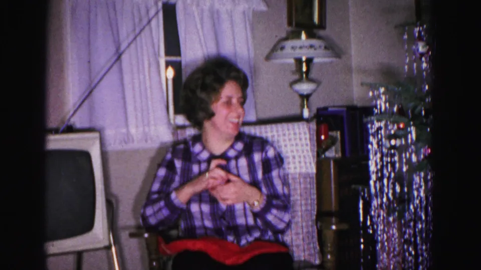 A woman in a purple plaid blouse holds a red gift item in a circa-1962 American living room; a boxy CRT television and hanging tinsel decoration are visible behind her.