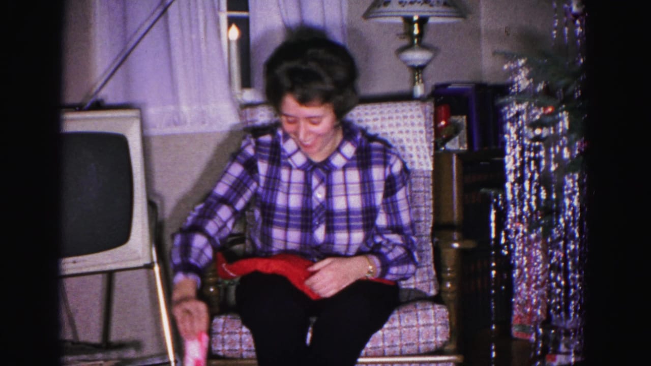 Woman in purple plaid blouse smiling over a red gift in a circa-1962 American living room; a hanging silver tinsel decoration and period CRT television are visible in the background.