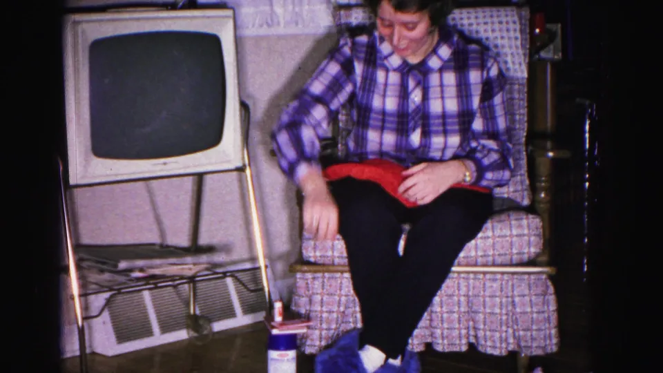 Woman examining a red gift in her lap in a circa-1962 American domestic interior; a boxy CRT television, patterned upholstery, and items on the floor are visible.