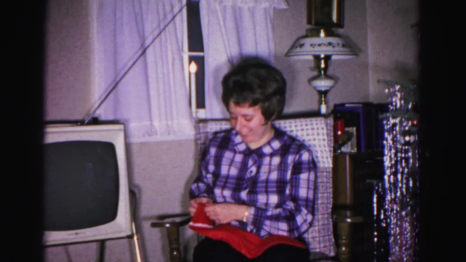 Woman in plaid blouse studying a red gift in a circa-1962 American living room; a decorative floor lamp, hanging tinsel decoration, and period CRT television are visible behind her.