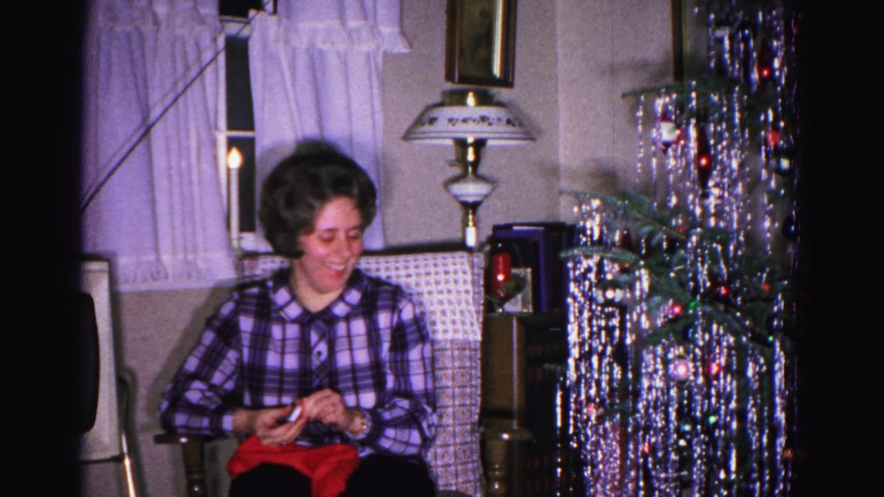 Woman in plaid shirt looks down handling a red object, with a dense tinsel Christmas tree and decorative lamp visible behind her.