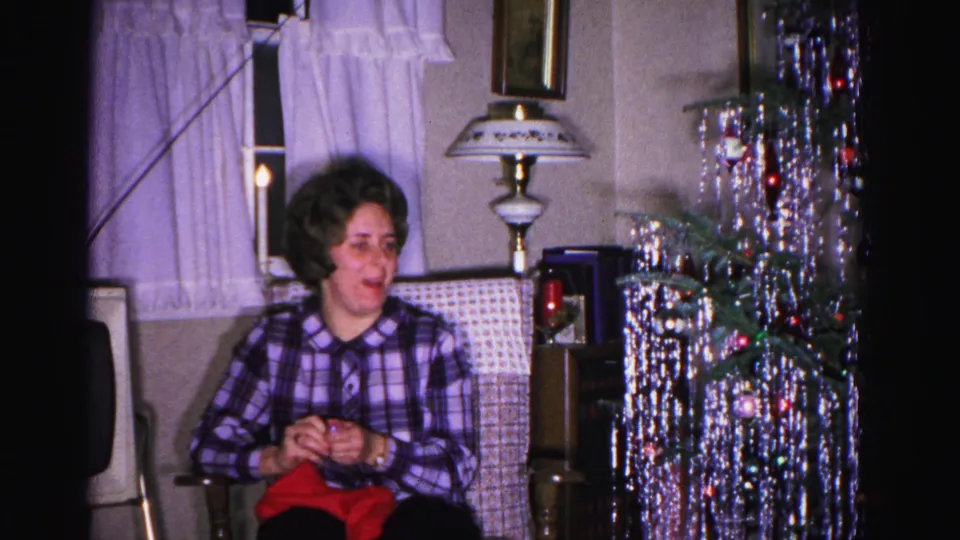 Woman in purple plaid shirt smiles beside a dense silver tinsel Christmas tree with colored ornaments and a tall decorative table lamp, Hagerstown MD, 1962.