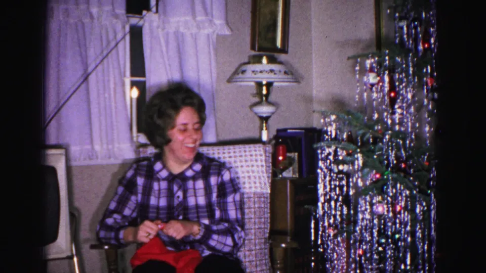 Woman in purple plaid shirt smiles down at a red object in her lap beside a tinsel-adorned Christmas tree and decorative lamp in a 1962 Hagerstown MD home.