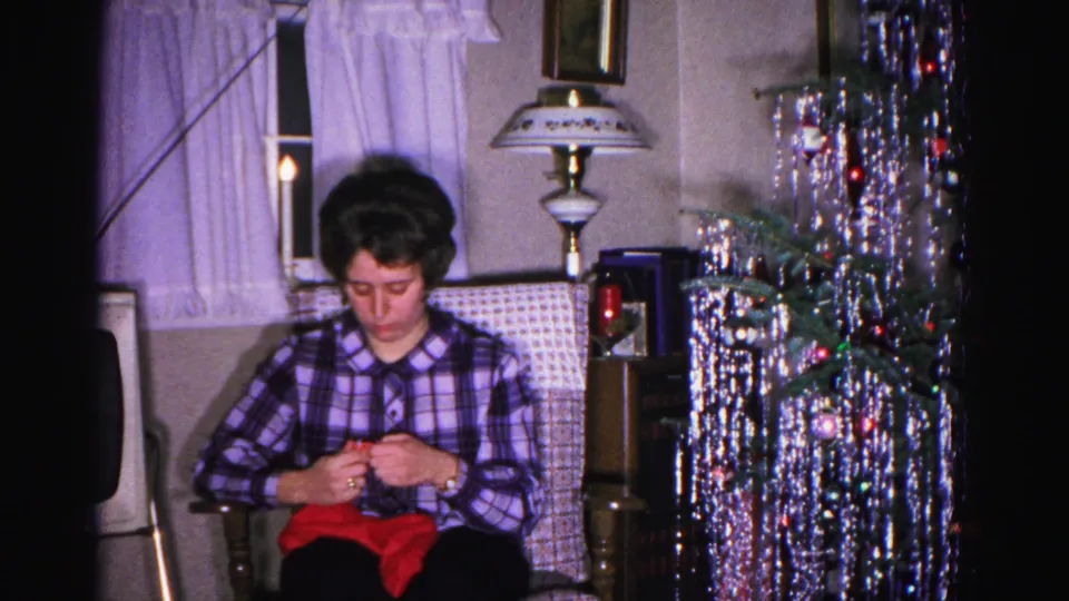 Medium-close shot of woman knitting in armchair, silver tinsel tree at right edge, curtains behind; Hagerstown, Maryland, ca. 1962.