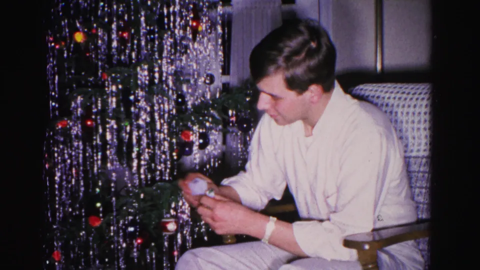 Young man examining or placing a ball ornament beside a tinsel-draped Christmas tree in a mid-century American home, Hagerstown, Maryland, circa 1962.